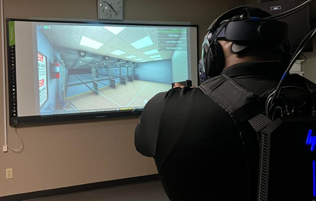 A person wearing a VR headset equipped with a tactical vest and VR gear is engaged in a virtual training simulation using the Apex Officer system. A large screen in the room displays a virtual shooting range environment with options such as "Start range" and "Next range mode." A clock is mounted on the wall above the screen.