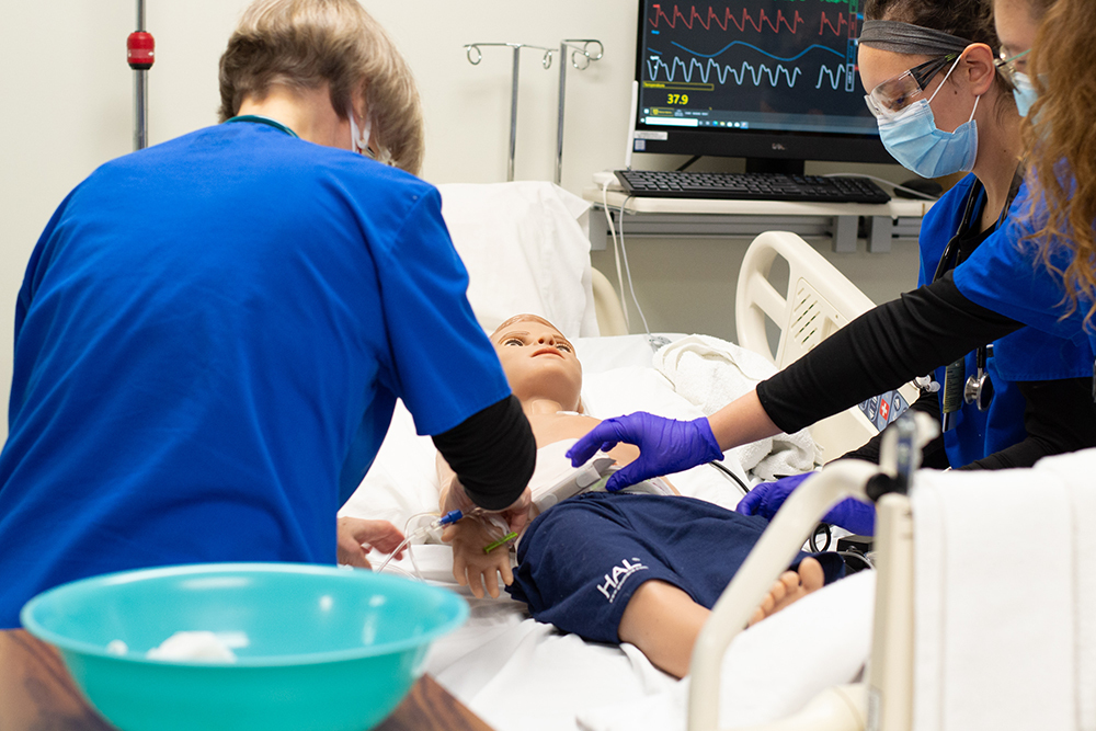 Medical trainees in blue scrubs perform a simulated procedure on a pediatric mannequin in a hospital bed, with monitoring equipment displaying vital signs in the background.