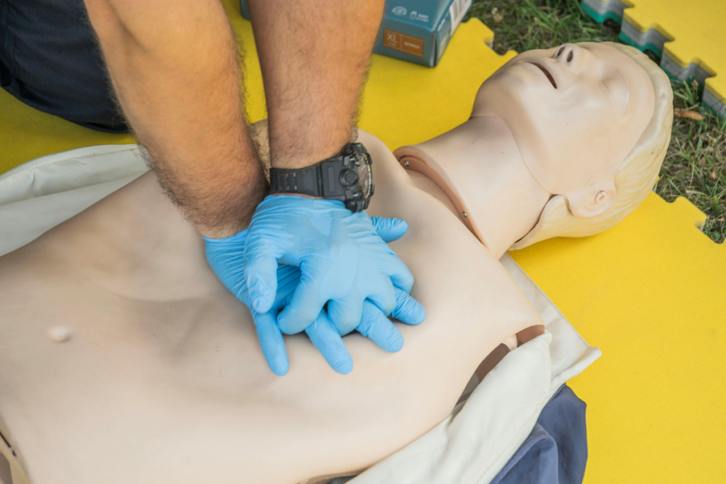 Hands wearing blue gloves performing CPR on a high-fidelity medical training mannequin placed on a yellow mat.