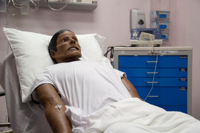 Simulation manikin lying in a hospital bed wearing a white shirt, used for clinical skills training at Edward Via College of Osteopathic Medicine.