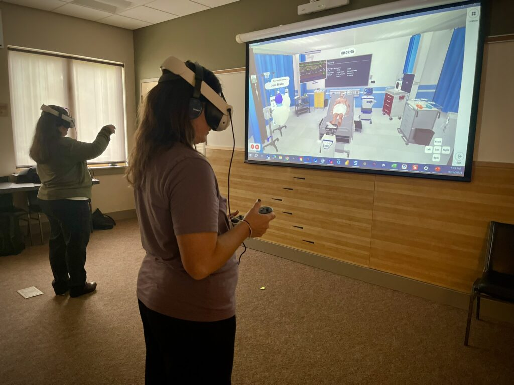 Two healthcare professionals wearing VR headsets and controllers participate in a Lumeto InvolveXR simulation, while the virtual scenario is displayed on a large screen in a classroom.