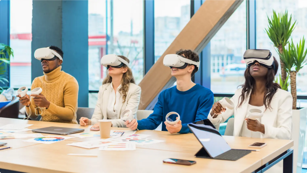 A diverse group of four individuals sits at a table wearing VR headsets and holding controllers, fully engaged in a virtual training session. The table is scattered with documents and laptops, suggesting a professional or learning environment.